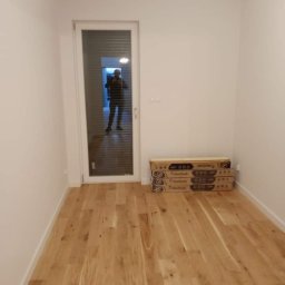 Emilian Fedorowicz Usługi remontowo budowlane - Jasne panele podłogowe w nowym pomieszczeniu z białymi ścianami, widoczne pudełka z panelami przy ścianie i odbicie osoby w przeszklonych drzwiach.