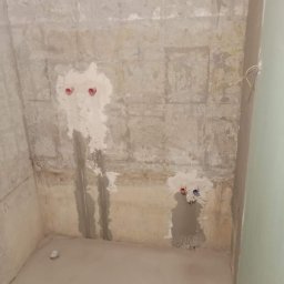 Emilian Fedorowicz Usługi remontowo budowlane - Surowa ściana w trakcie przygotowania do wykończenia, widoczne ślady po instalacjach wodnych i elektrycznych, nierówna powierzchnia oraz świeża wylewka na podłodze.