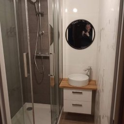 Emilian Fedorowicz Usługi remontowo budowlane - Nowoczesna kabina prysznicowa z szarymi płytkami, okrągłe lustro z odbiciem osoby robiącej zdjęcie, biała umywalka nablatowa na drewnianym blacie i podwieszana szafka z dwiema szufladami, jasne...