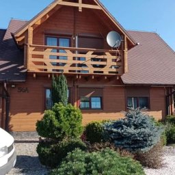 Emilian Fedorowicz Usługi remontowo budowlane - Drewniany dom jednorodzinny z balkonem, brązowy dach, satelita na elewacji, zadbany ogród z iglakami, biały samochód dostawczy częściowo widoczny z lewej strony.