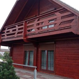 Emilian Fedorowicz Usługi remontowo budowlane - Czerwony, drewniany dom z balkonem, widok z zewnątrz, z widocznymi oknami z roletami i fragmentem chodnika z kostki.