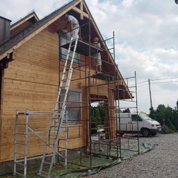 Emilian Fedorowicz Usługi remontowo budowlane - Dwóch mężczyzn na rusztowaniu maluje drewniany dom na zewnątrz, widoczna drabina, zabezpieczone okno folią i biały van w tle.