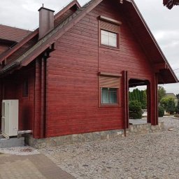 Emilian Fedorowicz Usługi remontowo budowlane - Czerwony drewniany dom z brązowymi roletami zewnętrznymi, widok z boku na elewację i podjazd z kostki brukowej, klimatyzator na zewnątrz.