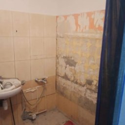 Emilian Fedorowicz Usługi remontowo budowlane - Zdemontowane płytki w łazience, odsłonięta warstwa starego tynku, widoczna umywalka i fragment rury, przygotowanie do dalszych prac.