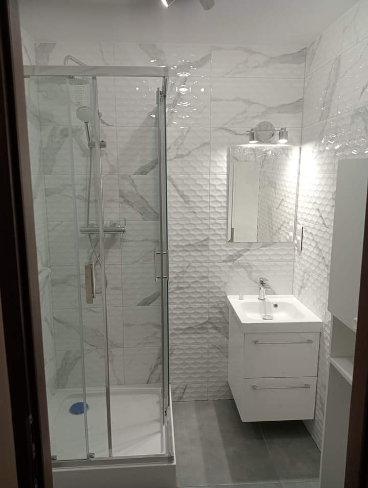 Nowoczesna kabina prysznicowa z białymi płytkami imitującymi marmur i umywalką z szafką w wykończonej łazience w Kaliszu, widoczne oświetlenie punktowe nad lustrem.