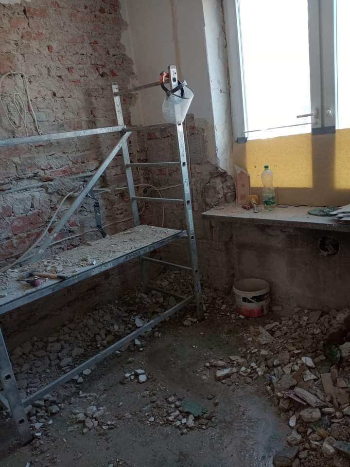 Wnętrze w trakcie demontażu starej okładziny ściennej, widoczne rusztowanie, gruz na podłodze i odsłonięta cegła, butelka wody mineralnej na parapecie okiennym.