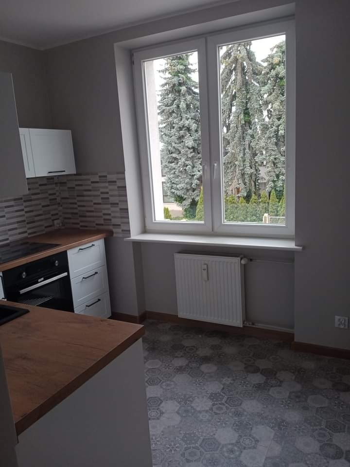 Nowoczesna kuchnia w szarościach z drewnianym blatem, wzorzystą podłogą i dużym oknem z widokiem na drzewa iglaste.