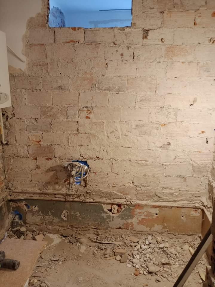 Ceglana ściana w trakcie skuwania tynku, widoczne przewody elektryczne w niebieskiej rurce, gruz na podłodze, fragment drabiny po prawej stronie.