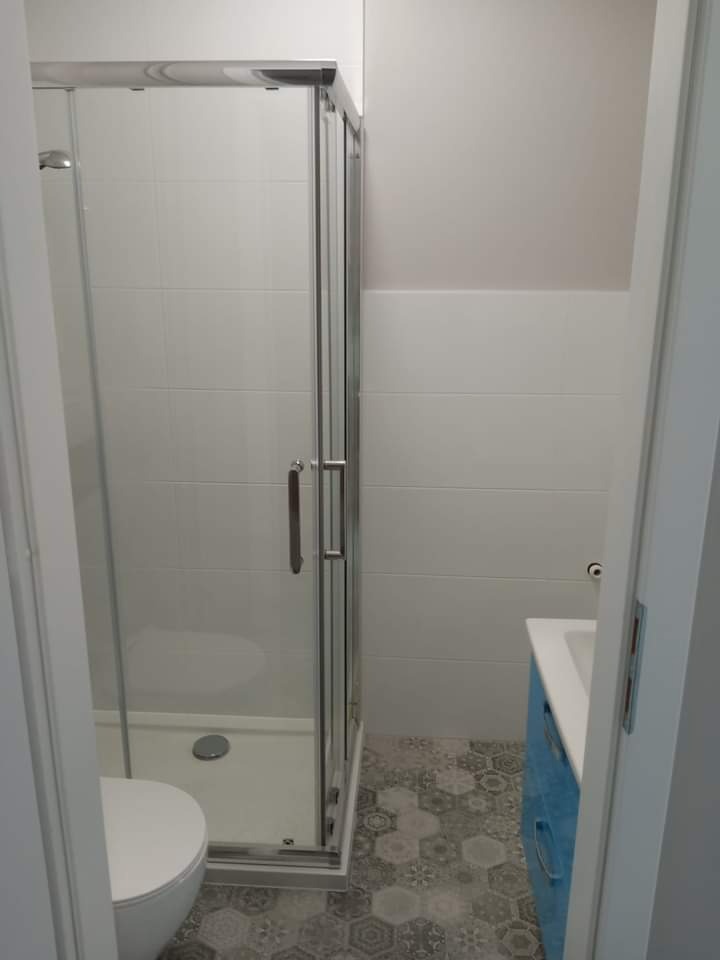 Wyremontowana łazienka z kabiną prysznicową z przesuwnymi drzwiami, toaletą, umywalką w niebieskiej szafce i podłogą wyłożoną szarymi heksagonalnymi płytkami.