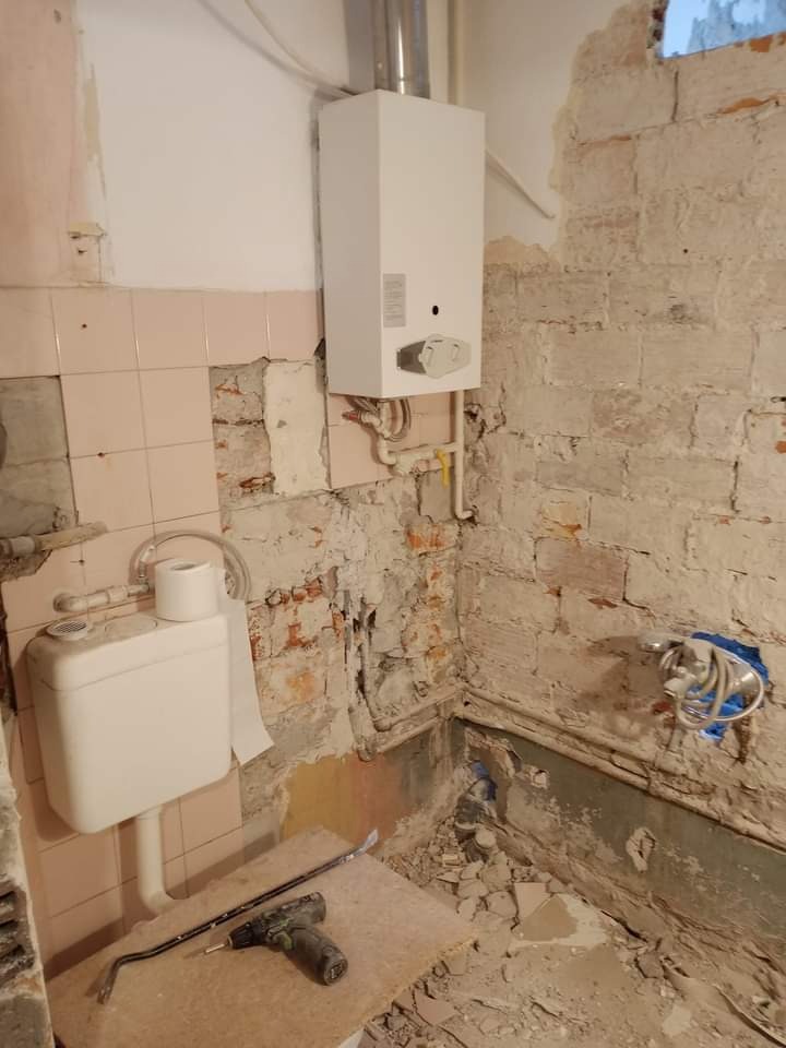 Demontaż starej instalacji w trakcie prac remontowych w łazience, widoczne fragmenty starych płytek, odsłonięta cegła, nowy piec gazowy, narzędzia i gruz.