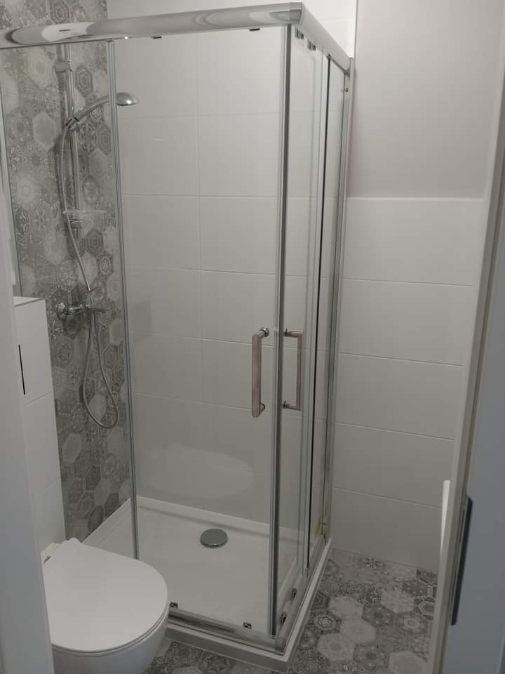 Nowoczesna kabina prysznicowa z chromowanymi profilami i szklanymi drzwiami, widoczna toaleta i płytki w geometryczne wzory na ścianie prysznica i podłodze.