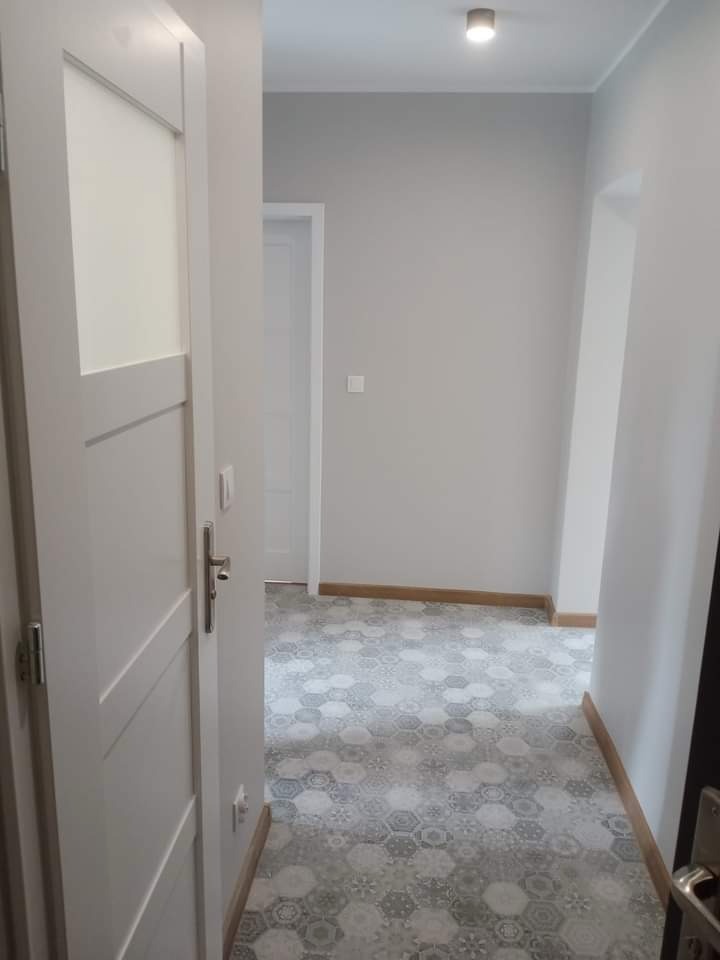 Korytarz po remoncie z szarymi ścianami, heksagonalną płytką podłogową w odcieniach szarości i bieli, oraz białymi drzwiami z przeszkleniem.