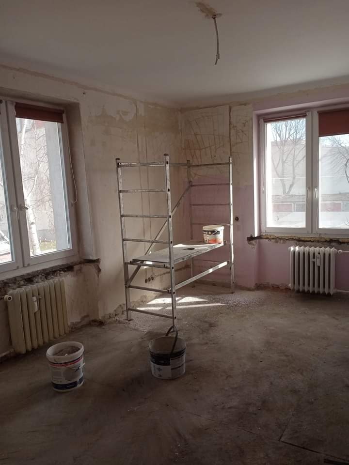 Pokój w trakcie przygotowań do prac wykończeniowych: ściany częściowo pokryte gładzią, aluminiowe rusztowanie, dwa wiadra z farbą na podłodze, odsłonięte grzejniki pod oknami.