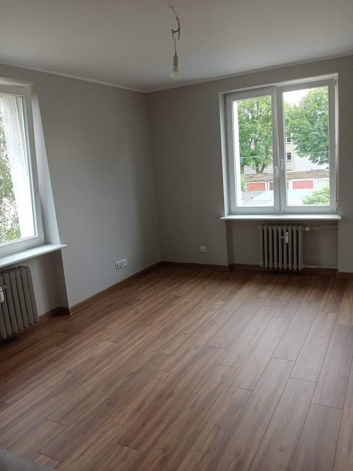 Pomalowane na szaro puste pomieszczenie z panelami podłogowymi w odcieniach brązu, dwa okna z białymi ramami i grzejnikami pod nimi, goła żarówka zwisająca z sufitu.