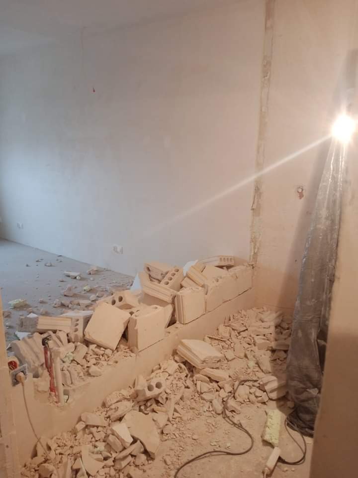 Demontaż fragmentu ściany z cegły w pomieszczeniu, widoczne gruz i odsłonięte instalacje elektryczne.