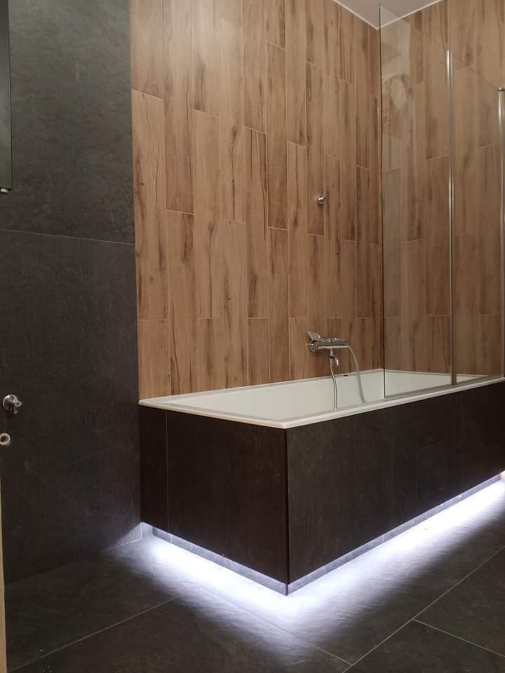 Wykończona łazienka z wanną obudowaną ciemnymi płytami imitującymi kamień, podświetloną listwą LED, drewnopodobnymi płytkami na ścianie i szklanym parawanem nawannowym.