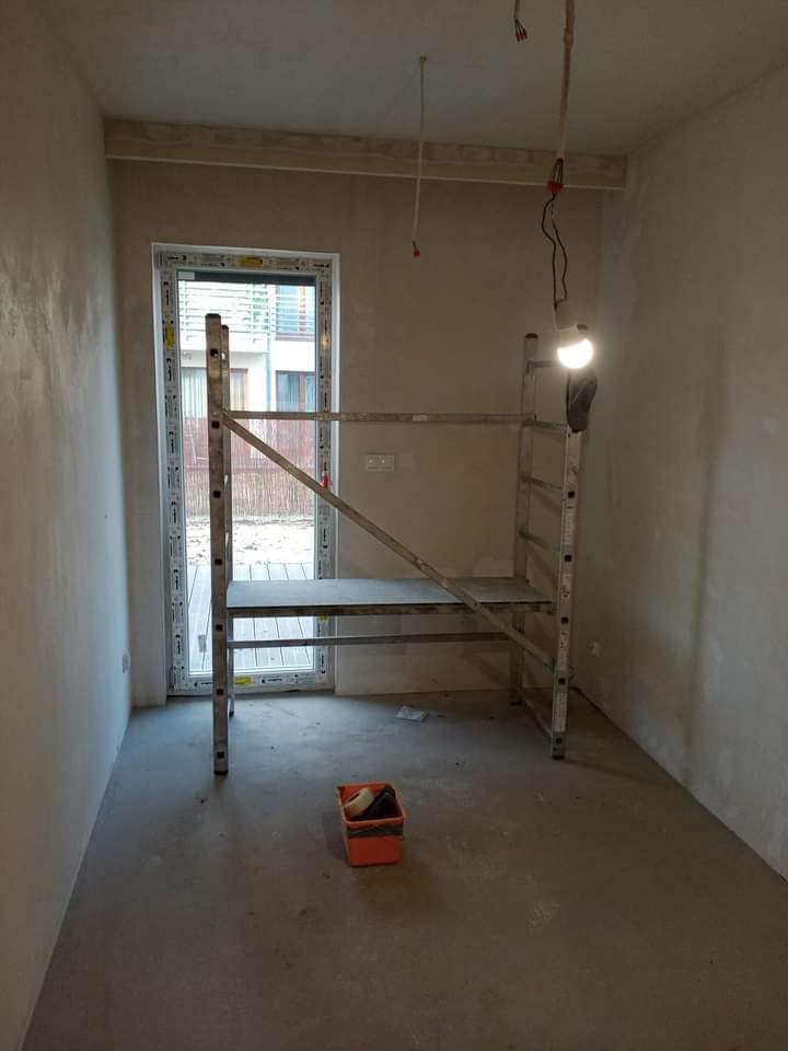 Pokój w trakcie prac wykończeniowych: ściany pokryte świeżo nałożoną warstwą gładzi, aluminiowe rusztowanie z lampą roboczą, pomarańczowe wiaderko z narzędziami malarskimi na podłodze, okno...