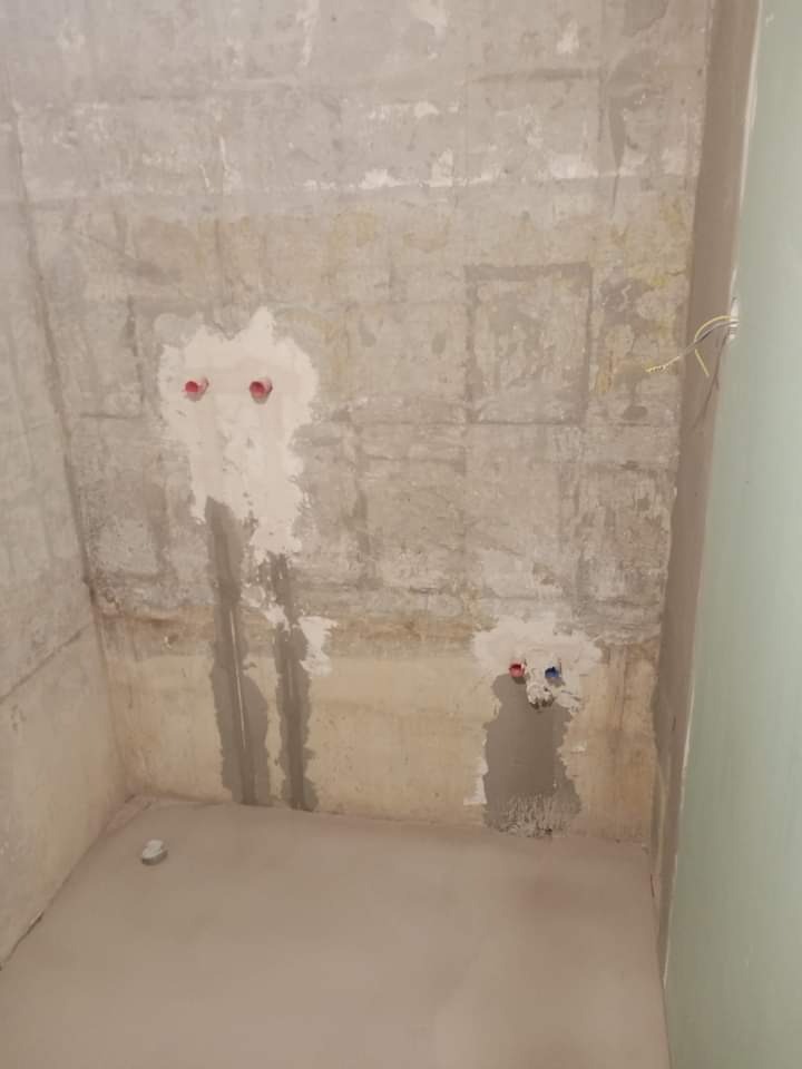 Surowa ściana w trakcie przygotowania do wykończenia, widoczne ślady po instalacjach wodnych i elektrycznych, nierówna powierzchnia oraz świeża wylewka na podłodze.