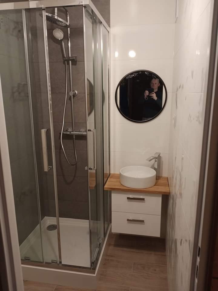 Nowoczesna kabina prysznicowa z szarymi płytkami, okrągłe lustro z odbiciem osoby robiącej zdjęcie, biała umywalka nablatowa na drewnianym blacie i podwieszana szafka z dwiema szufladami, jasne...