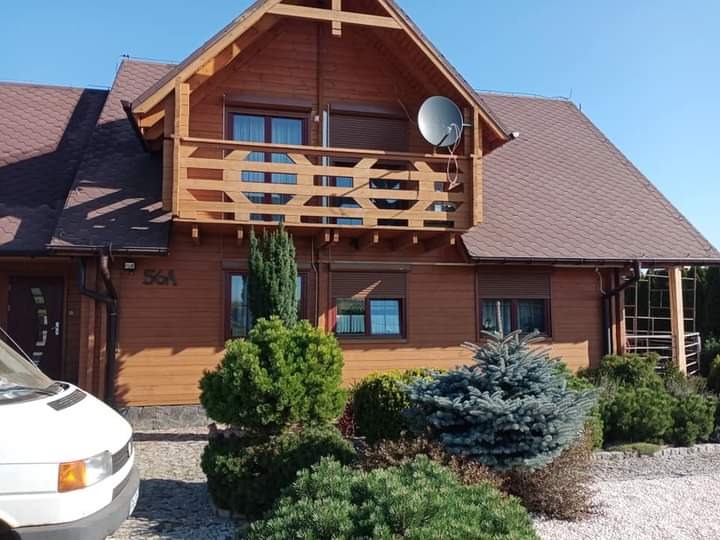 Drewniany dom jednorodzinny z balkonem, brązowy dach, satelita na elewacji, zadbany ogród z iglakami, biały samochód dostawczy częściowo widoczny z lewej strony.