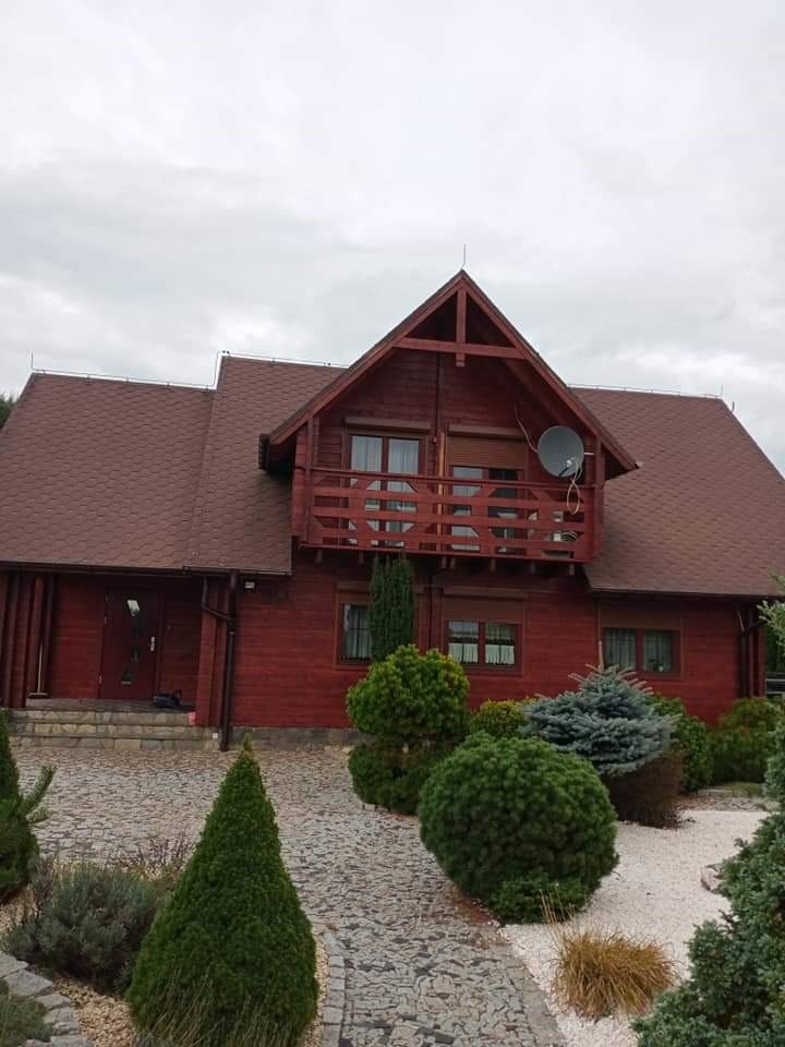 Dwupiętrowy dom z bali pomalowany na czerwono-brązowy kolor z brązową dachówką, balkonem z drewnianą balustradą i anteną satelitarną, otoczony ozdobnymi krzewami i kamienną ścieżką.