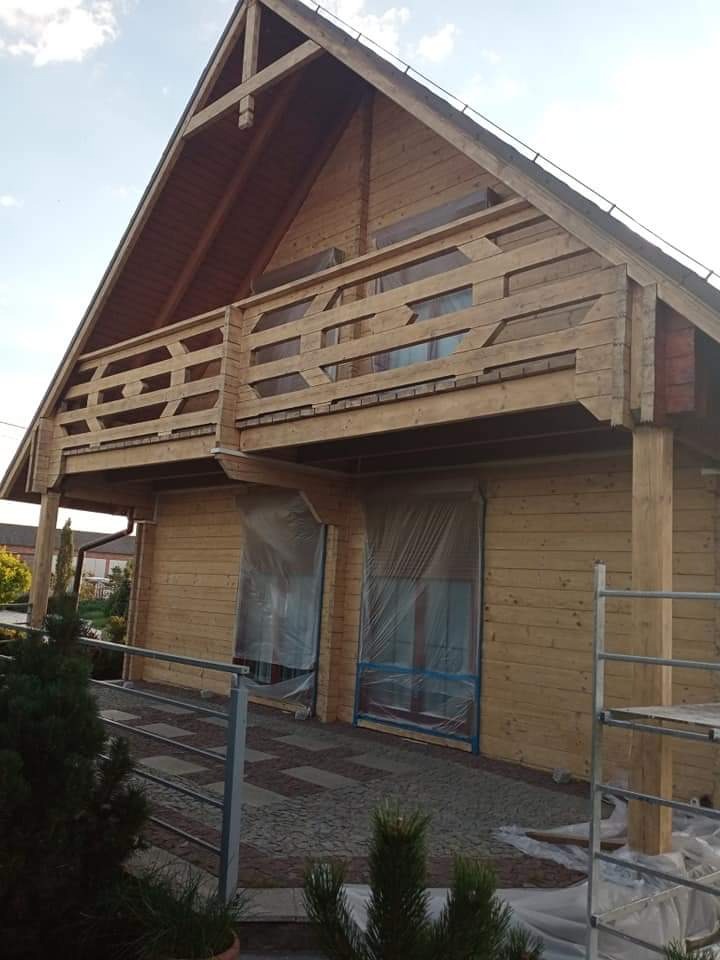 Drewniany dom z balkonem, zabezpieczony folią malarską, widoczny fragment rusztowania, świeżo pomalowana elewacja.