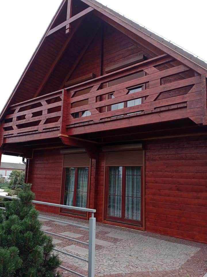 Czerwony, drewniany dom z balkonem, widok z zewnątrz, z widocznymi oknami z roletami i fragmentem chodnika z kostki.