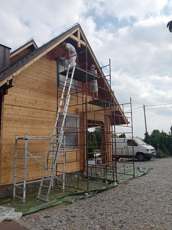 Dwóch mężczyzn na rusztowaniu maluje drewniany dom na zewnątrz, widoczna drabina, zabezpieczone okno folią i biały van w tle.