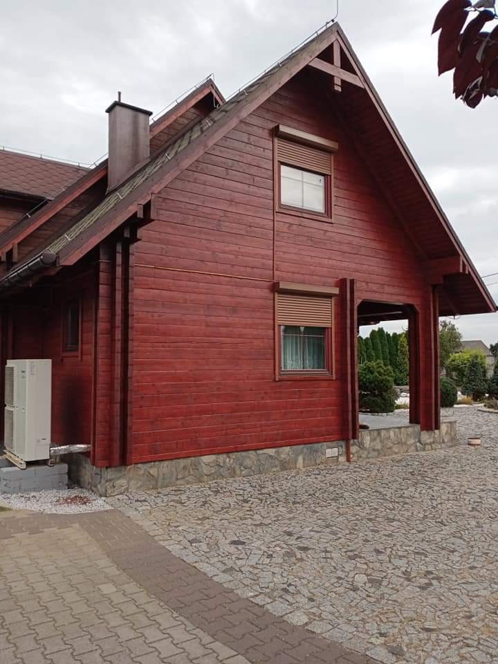 Czerwony drewniany dom z brązowymi roletami zewnętrznymi, widok z boku na elewację i podjazd z kostki brukowej, klimatyzator na zewnątrz.