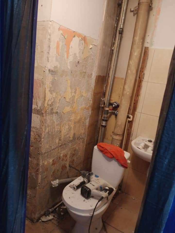 Zdemolowana łazienka w trakcie remontu: ściany z odpadającą farbą, rury, toaleta z narzędziami, widoczne ślady po kuciu.