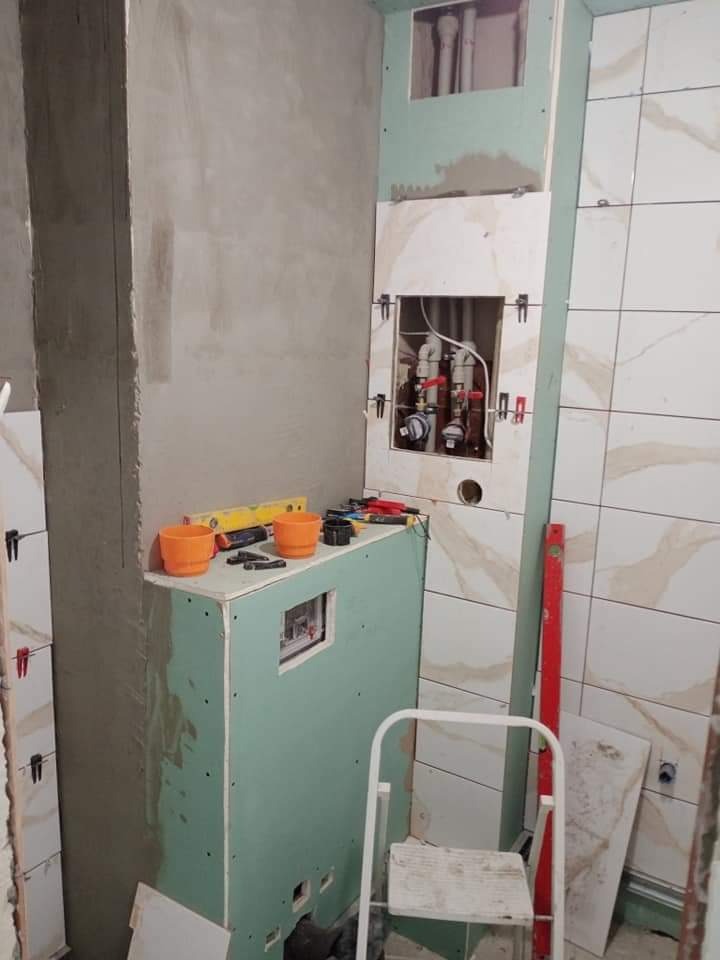 Łazienka w trakcie remontu: zabudowa z płyt kartonowo-gipsowych, widoczne rury i zawory, narzędzia na górze zabudowy, częściowo ułożone płytki na ścianie.