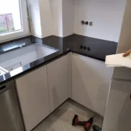 Montaż czarnego blatu kuchennego z widocznymi narzędziami (wiertarki) na podłodze, białe szafki kuchenne, widok na okno i niezabezpieczone kable w ścianie.