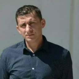 Portret mężczyzny w ciemnej koszuli na tle jasnej ściany, ujęcie od pasa w górę, z widocznym fragmentem górnej części ciała.