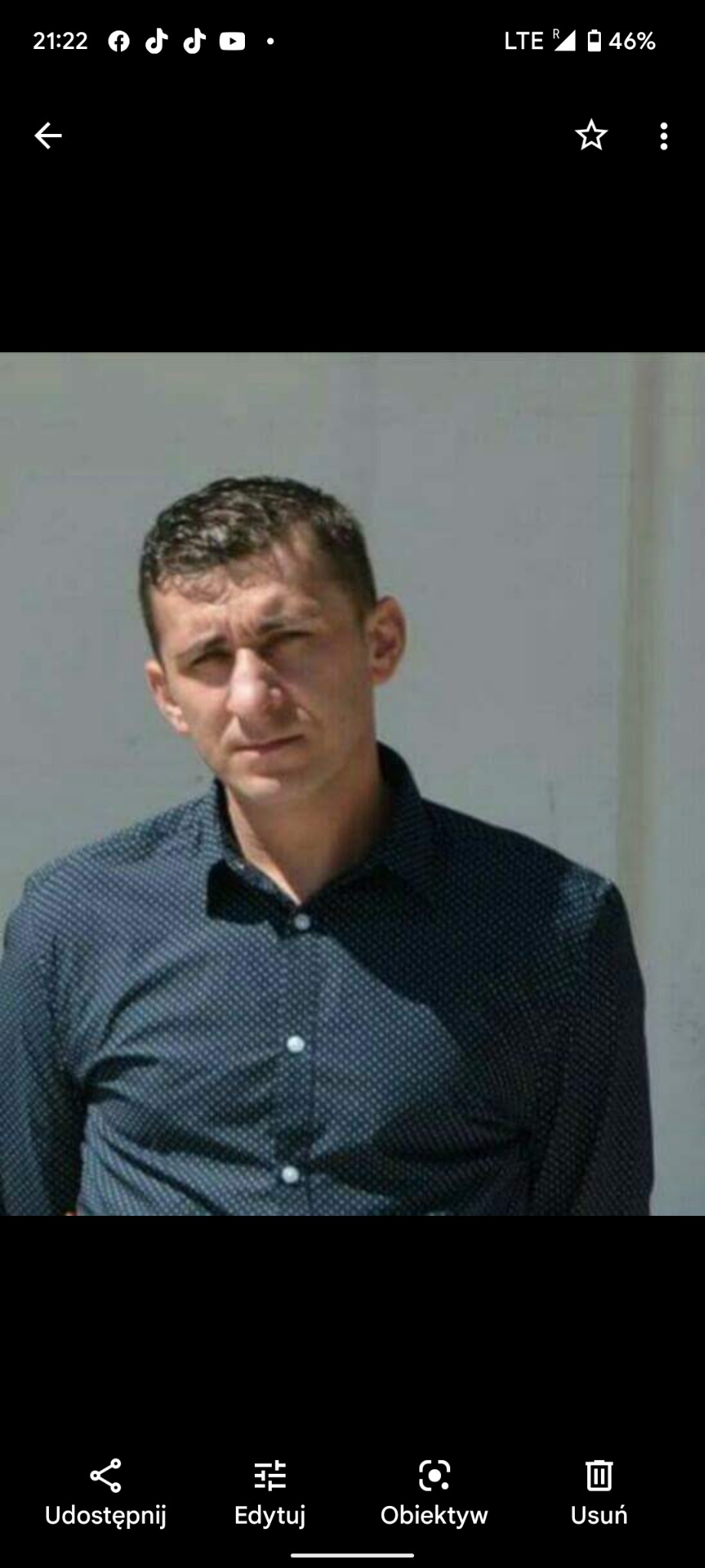 Portret mężczyzny w ciemnej koszuli na tle jasnej ściany, ujęcie od pasa w górę, z widocznym fragmentem górnej części ciała.