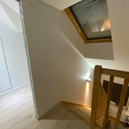 Wnętrze domu po remoncie: drewniane schody z balustradą, okno dachowe z widokiem na niebo, zamontowane oświetlenie punktowe na ścianie i jasna podłoga.