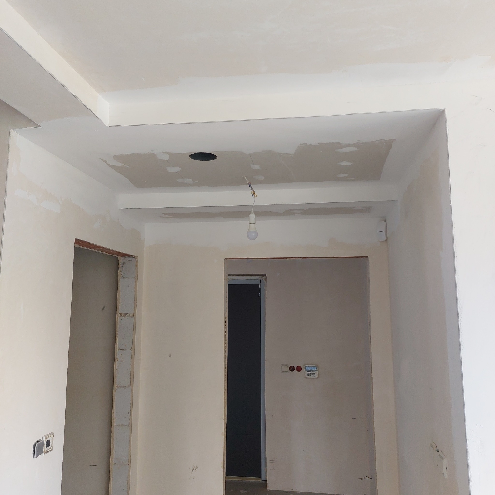 Wnętrze w trakcie prac wykończeniowych: sufit z elementami z płyty karton-gips, częściowo wyszpachlowany, widoczne otwory na oświetlenie, surowy stan ścian z widocznymi instalacjami elektrycznymi...