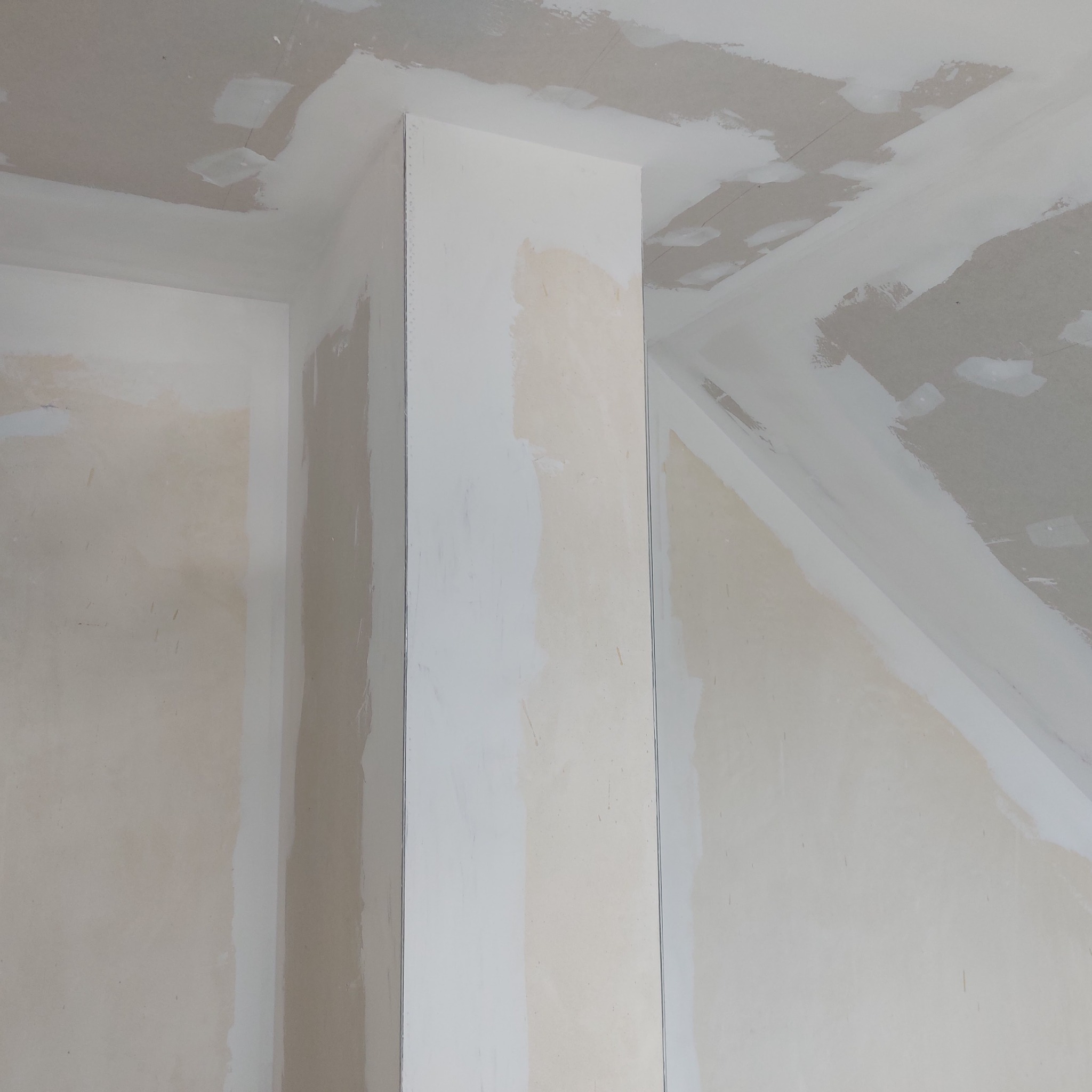 Wnętrze w trakcie prac wykończeniowych; widoczne ściany i sufit po nałożeniu warstwy szpachli, narożniki zabezpieczone listwami, sufit z widocznymi miejscami po łataniu.