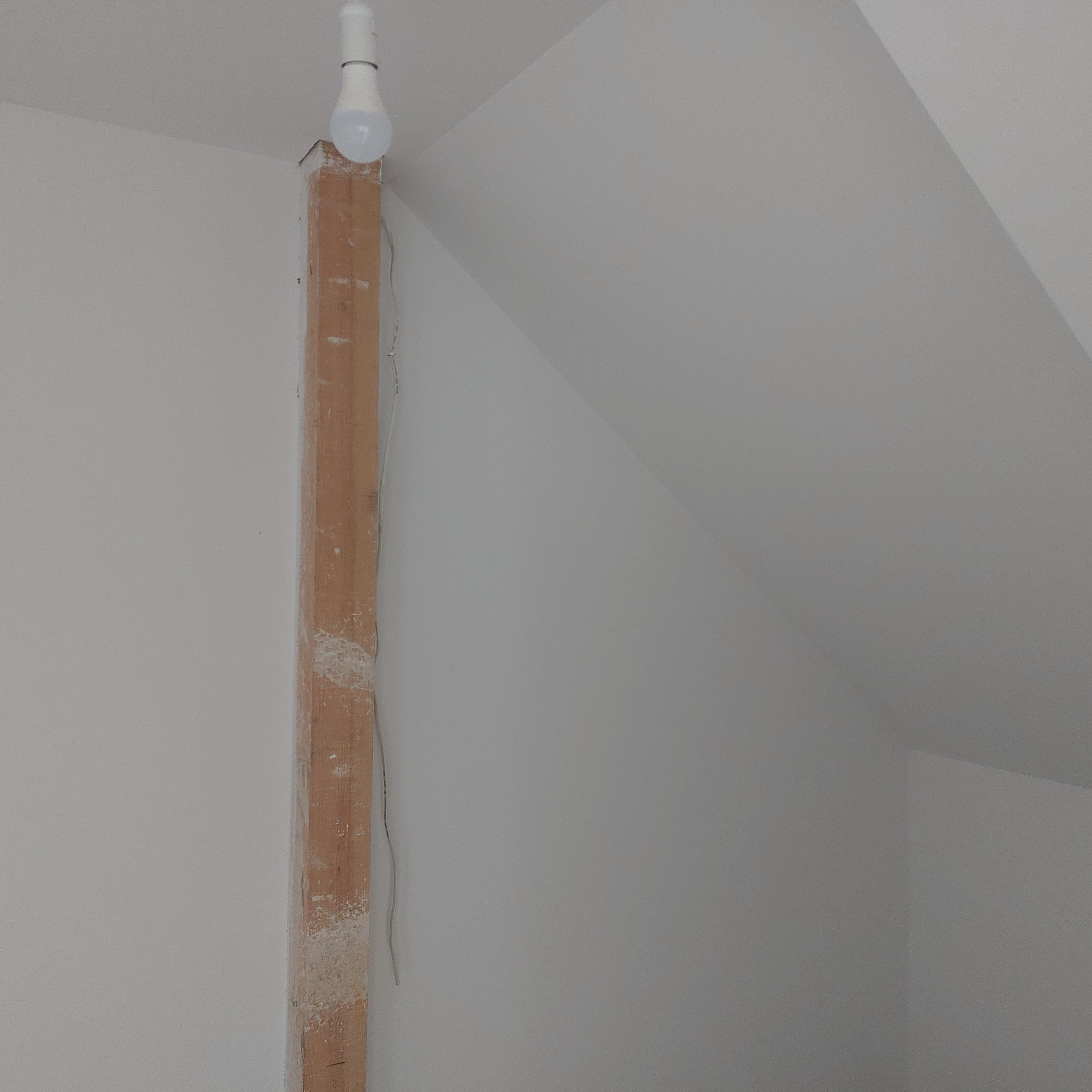 Wnętrze poddasza w trakcie prac wykończeniowych, widoczna surowa drewniana belka podtrzymująca z żarówką tymczasową, ściany przygotowane pod malowanie.