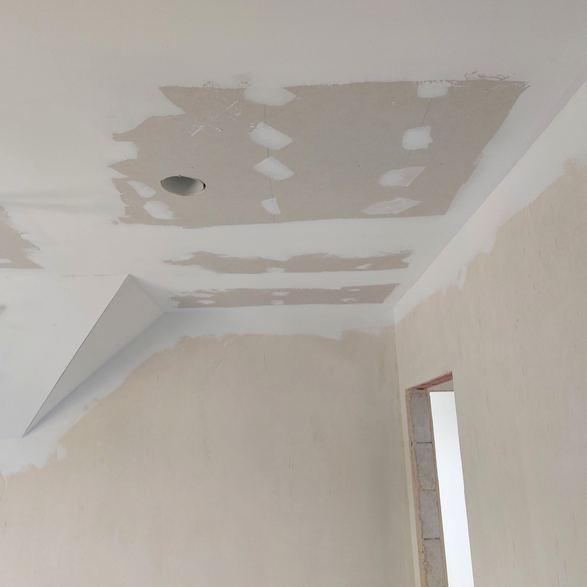 Wnętrze w trakcie prac wykończeniowych: fragment sufitu z widocznymi łatami szpachli, otworem na oświetlenie i fragmentem ściany z widocznymi śladami po pracach przygotowawczych.