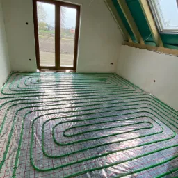 Zielone rurki ogrzewania podłogowego ułożone spiralnie na folii z czerwoną siatką w surowym pokoju na poddaszu z oknem dachowym i oknem balkonowym.