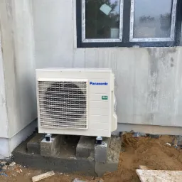 Zewnętrzna jednostka klimatyzacji Panasonic R32 zamontowana na betonowych podstawach przy ścianie budynku, widoczne okno z czarną ramą i surowy stan elewacji.