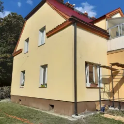 Żółty dom jednorodzinny z czerwoną dachówką po renowacji elewacji, widoczne okna z kwiatami, rynna, fragment rusztowania i pomarańczowy wąż ogrodowy na trawniku.