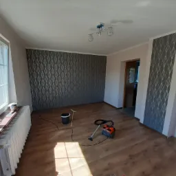 Pokój po remoncie z nową podłogą panelową, wzorzystą tapetą na ścianach, grzejnikiem pod oknem i sprzętem do sprzątania.