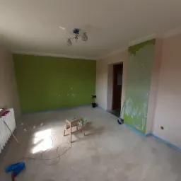 Pokój w trakcie malowania: jedna ściana zielona, druga różowa, widoczny stołek malarski, pędzel przy grzejniku i kabel na podłodze.