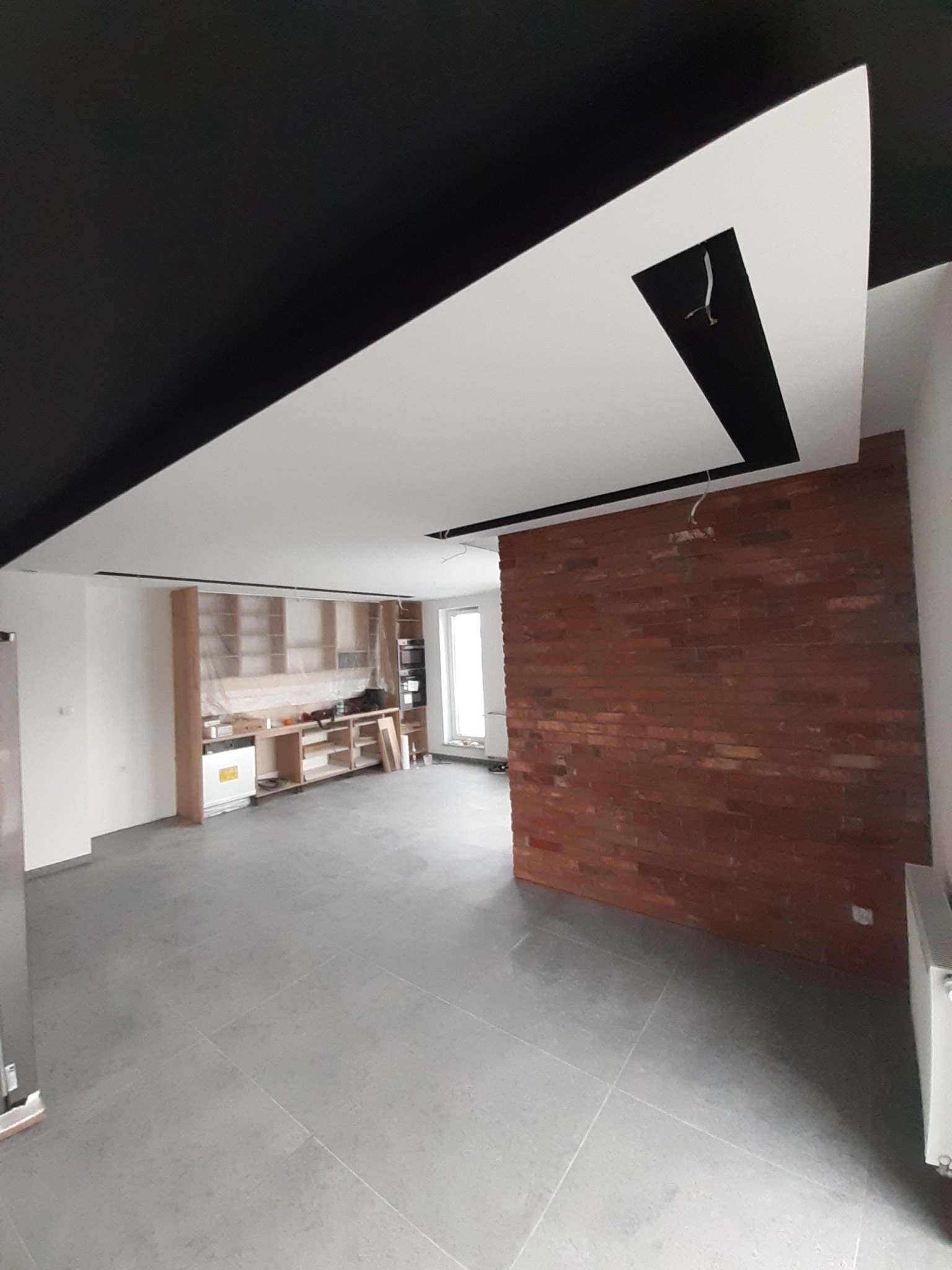 Wnętrze w trakcie wykończenia: szara podłoga z płytek, ściana z czerwonej cegły, czarny sufit z białym fragmentem z wycięciem na oświetlenie, zabudowa kuchenna częściowo zakryta folią ochronną.