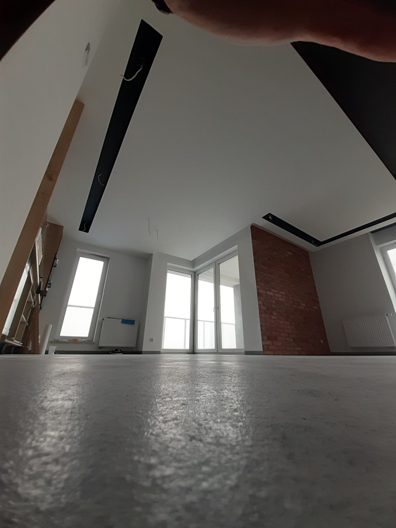 Wnętrze w trakcie wykańczania: widok z poziomu podłogi na sufit z czarnymi listwami, fragment ściany z cegły, okna i grzejniki, widoczne kable wystające z sufitu.