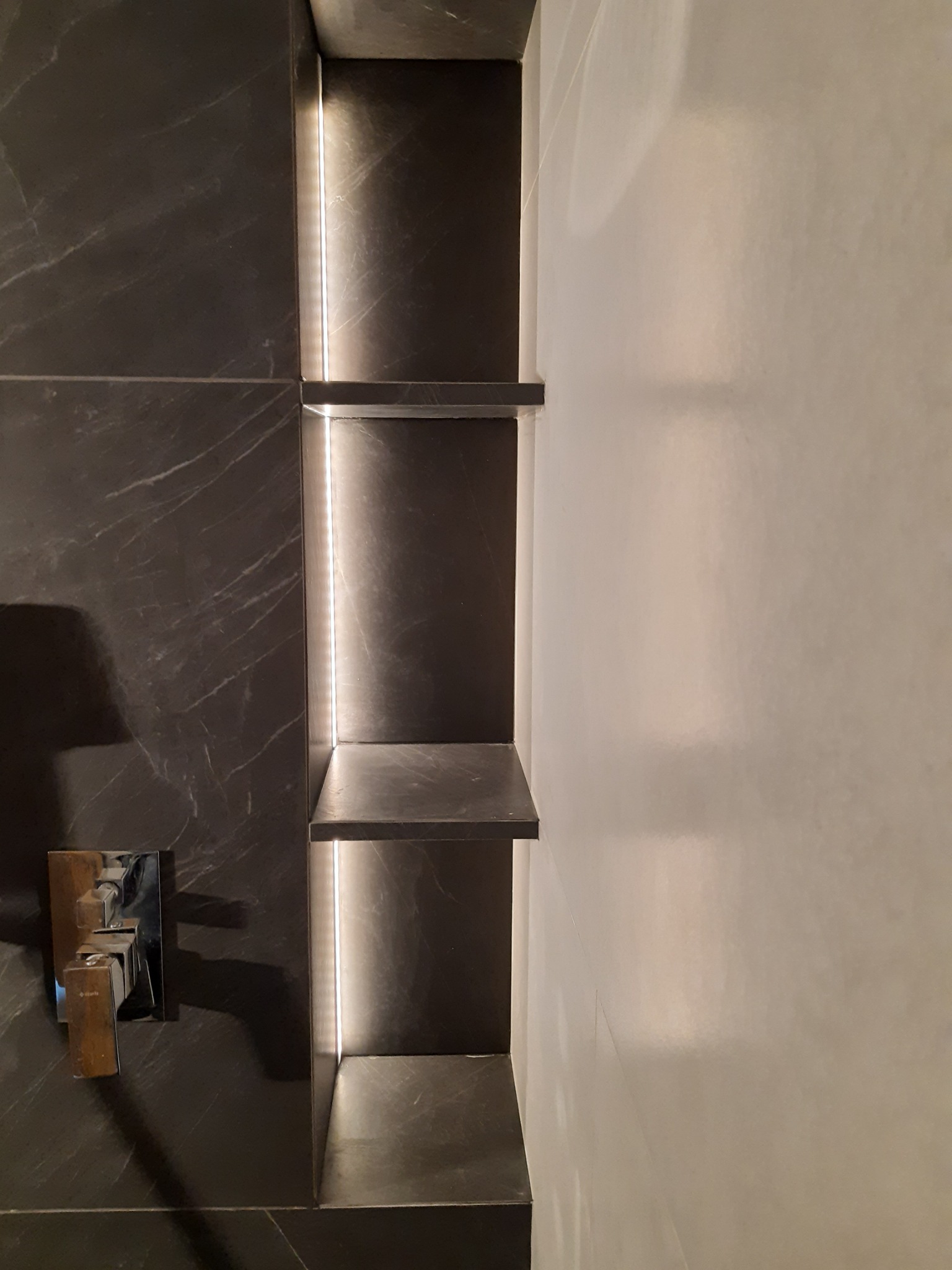 Nowoczesna łazienka z wnęką półkową podświetloną ledami, wykończona czarnym marmurem i jasnymi płytkami, widoczna armatura łazienkowa.