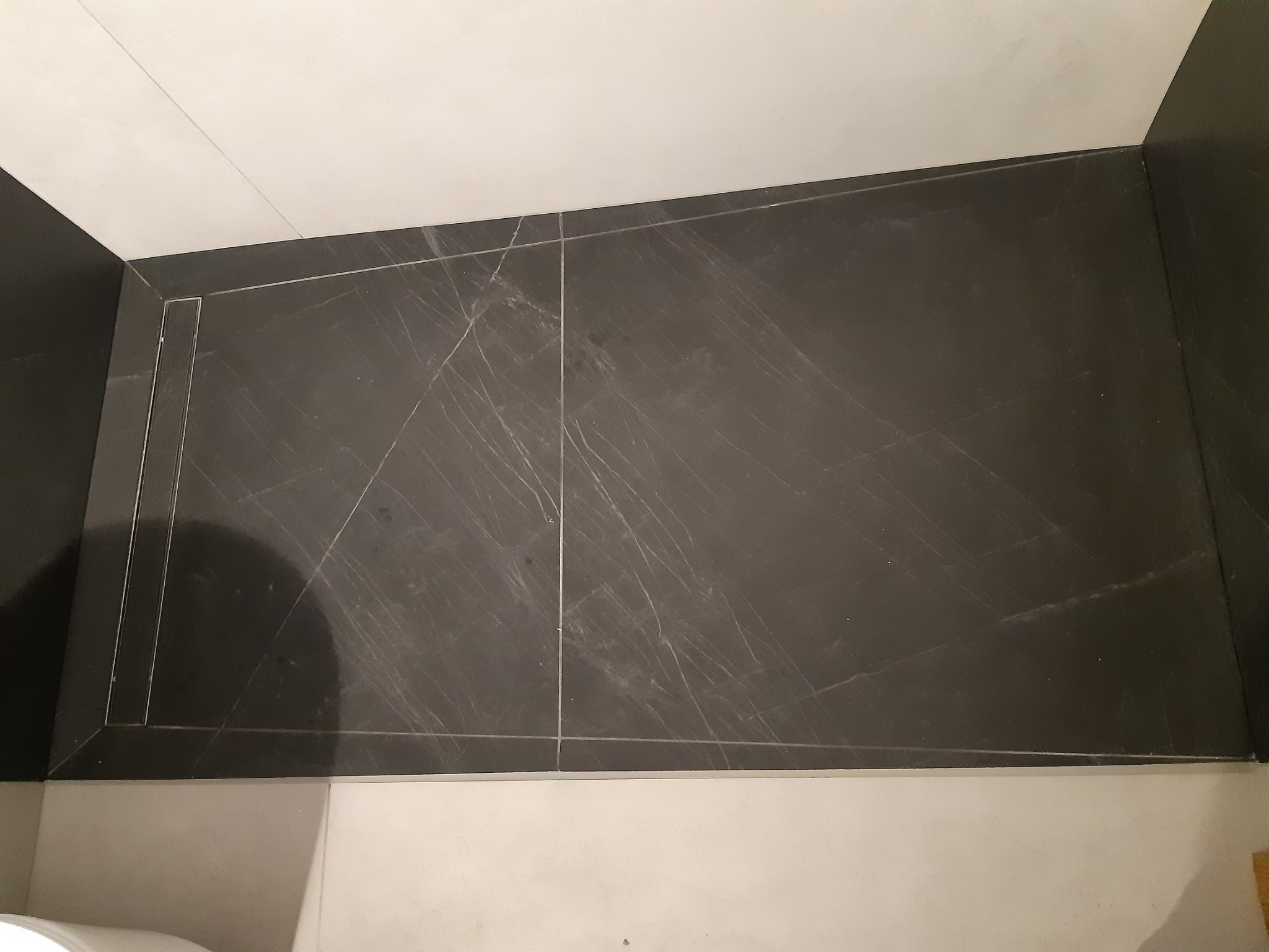 Wykończony brodzik z odpływem liniowym, wyłożony ciemnymi płytkami imitującymi marmur, w łazience w Olecku. Widoczne połączenie płytek podłogowych i ściennych.