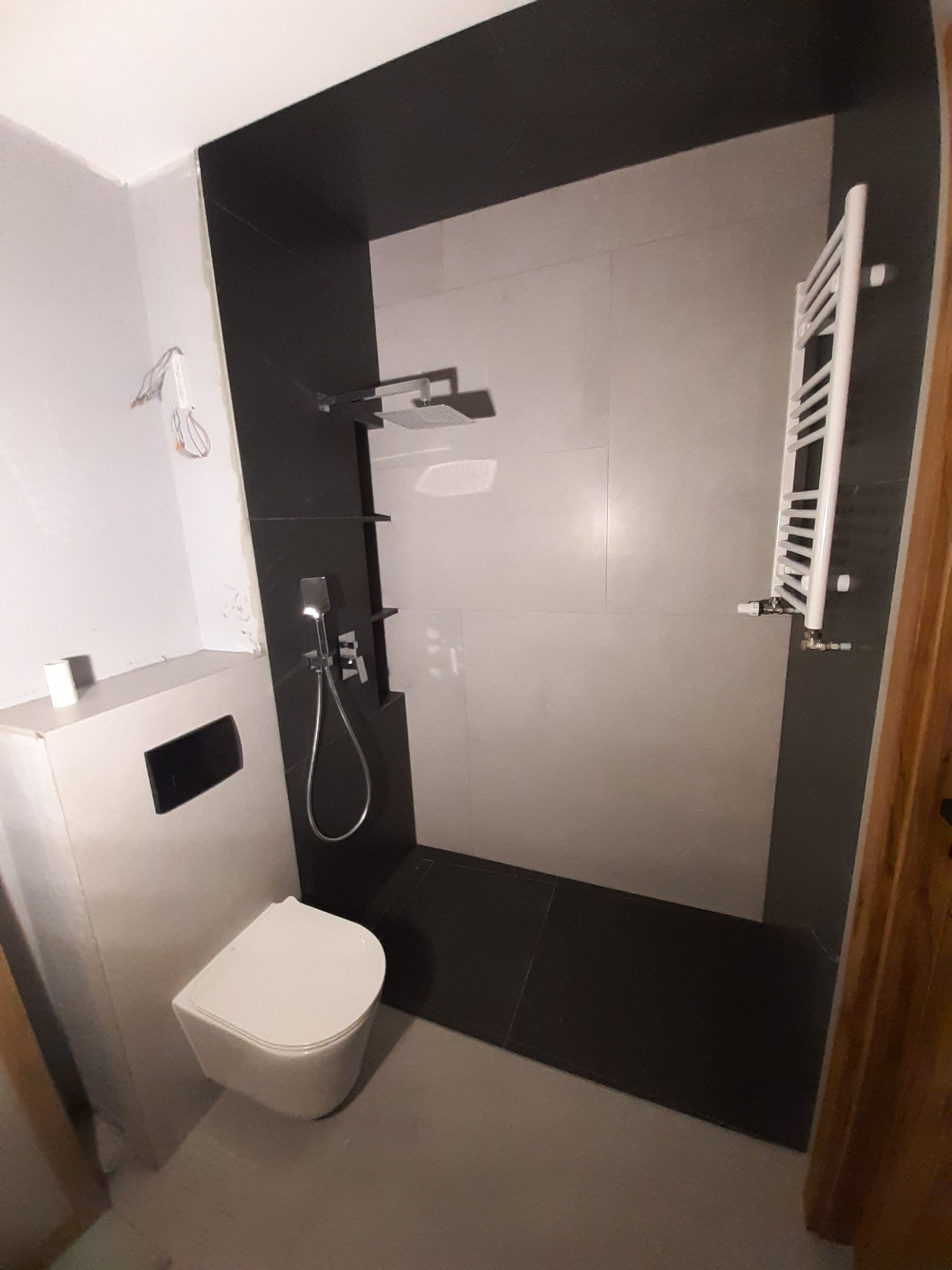Nowoczesna łazienka z kabiną prysznicową typu walk-in, podwieszaną toaletą i grzejnikiem drabinkowym, wykończona w szaro-czarnej kolorystyce z dużymi płytami.