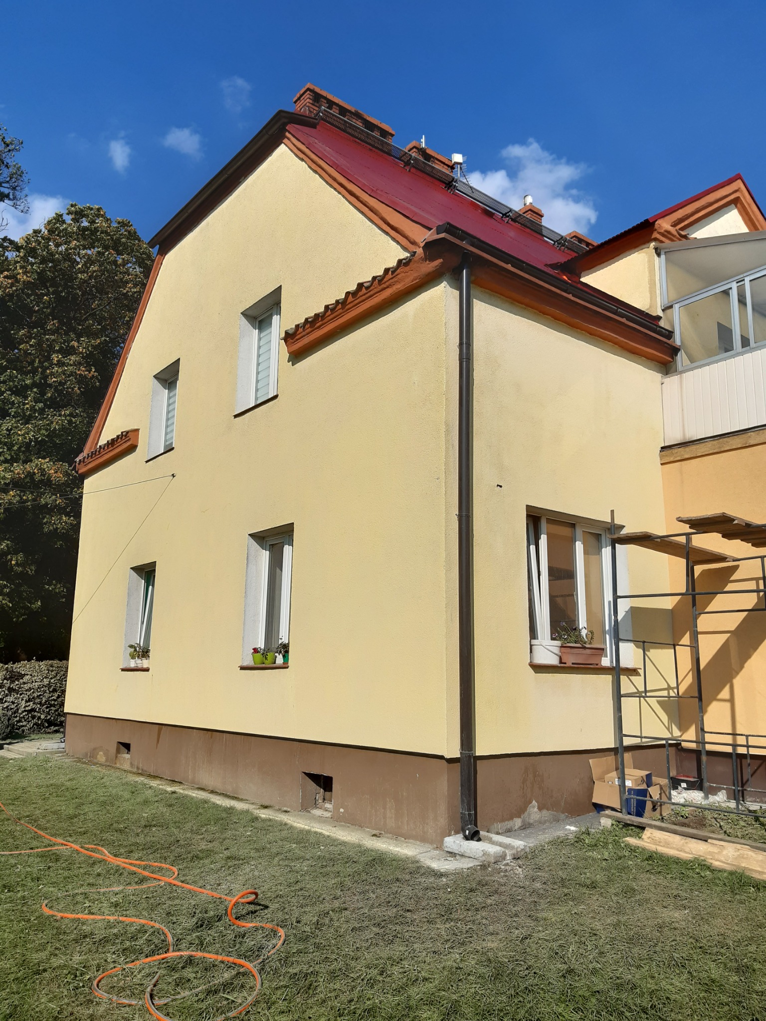Żółty dom jednorodzinny z czerwoną dachówką po renowacji elewacji, widoczne okna z kwiatami, rynna, fragment rusztowania i pomarańczowy wąż ogrodowy na trawniku.